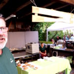 Links der Küchenmeister in einem grünen Shirt, hinter ihm die Küche und Essensausgabe mit dem Küchenteam