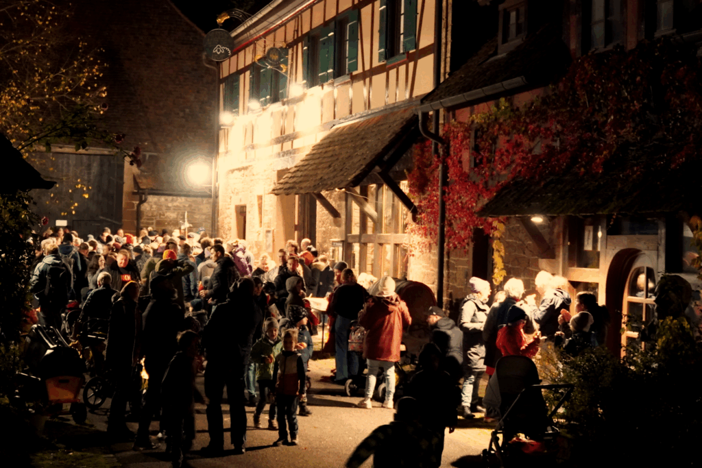 Bild vom Alt Maulbronner Laternlauf 2025. Das Bild wurdeauf dem Schafhof vor dem Museum aufgenommen. Es ist eine große Menschenmenge die auf dem Platz vor dem Museum in kleinen Gruppen steht. Vereinzelt sieht man Kinder mit Laternen und Leiterwagen. Es ist bereits dunkel und die Lampen tauchen das ganze Bild in ein warmes Licht