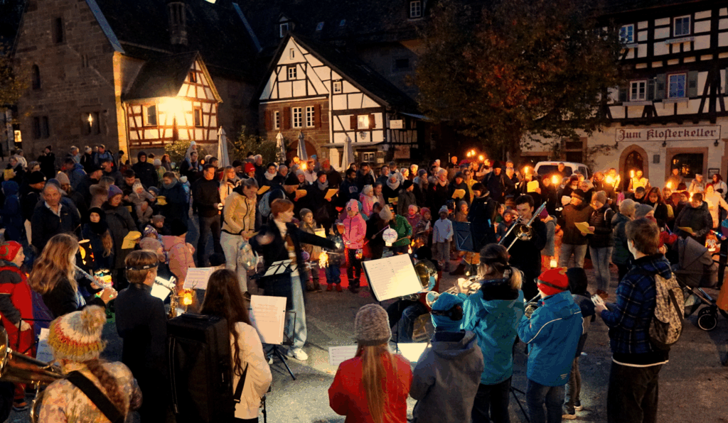 Bild vom Alt Maulbronner Laternlauf 2025. Das Bild wurde im Klosterhof aufgenommen, im Hintergrund sieht man Fachwerk und Sandsteingebäude des Klosterhofs. eine Menschenmenge aus Erwachsenen und kleinen Kindern mit Laternen ist über die komplette Breite Sichtbar. Mittig sieht man die Dirigentin des Schülerorchesters, die Musiker schauen in Richtung der Dirigentin und man sieht sie daher nur von hinten. Es dämmert bereits und die Lampen und Beleuchtung tauchen die Szenerie in ein warmes Licht