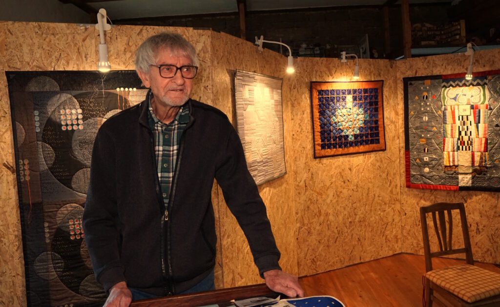 Das Bild zeigt Horst Keller, ein hagerer Mann mit grauem Haar und einem grauen Kinnbart. Er trägt eine schwarzrandige Brille, eine dunkle Strickjacke und darunter ein kariertes Hemd. Er hält gerade die Eröffnungsrede zur Ausstellung. Im Hintergrund sieht man an einer Holzwandel verschiedene aufgehängte Quilts.