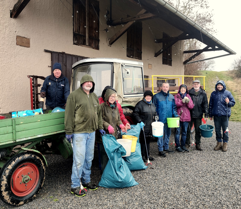 Ein Gruppenbild von 8 Personen. Sie stehen in einer Reihe vor einem landwirtschaftlichen Anhänger und Traktor. Sie haben Eimer und blaue Mülltüten in der Hand. Zum Zeitpunkt der Aufnahme war es regnerisch, daher tragen alle Personen Jacken und teilweise Mützen. Trotz allem sehen Sie zufrieden aus.