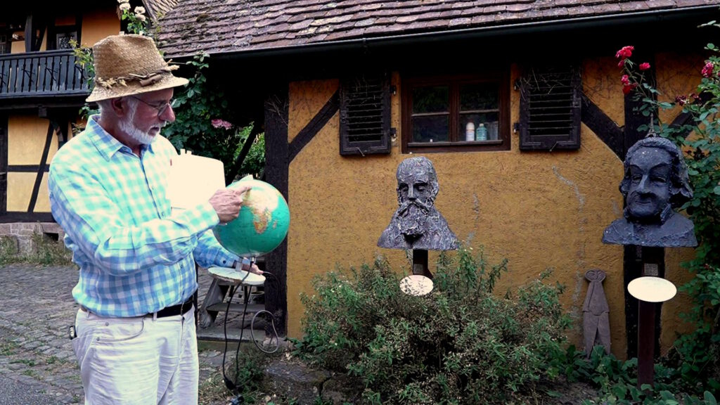 Links im Bild ein Mann mit Strohhut und karierten Hemd. Er hat einen weißen Kinnbart. Er hält einen Globus in der Hand und zeigt mit seiner rechten Hand darauf. In der Mitte und rechts sind zwei Büsten zu sehen. Die Mittlere Büste ist die von Ferdinand von Hochstetter. Die Büste rechts ist von Karl Friedrich Reinhard